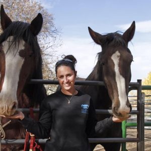 Margaret Blaha leaning against a fence. Behind the fence are two brown horses. She is petting their noses.