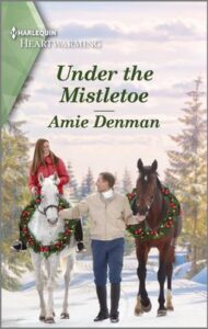 Cover image for Under the Mistletoe by Amie Denman, featuring a man guiding two horses down a snowy path. The horse on the left is white and has a woman riding it. The horse on the right is brown. Both horses are wearing Christmas wreaths around their necks.