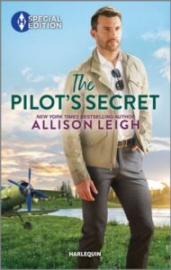 Cover image for The Pilot's Secret by Allison Leigh, featuring a man standing by a lake. He is wearing a jacket and has aviator sunglasses hooked into the collar of his shirt. Behind him next to the lake is a small airplane with a propeller in the front.