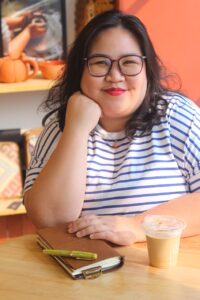Image of author Carla de Guzman, sitting at a table with a notebook, a pen and an iced coffee. Her hand is resting in her hand and she is smiling at the viewer. 