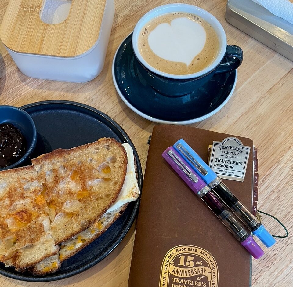 Image of a table top that includes a plate with a Kesong Puti Grilled Cheese Sandwich, a latte with a heart made in the foam, and a notebook with multicoloured pens.