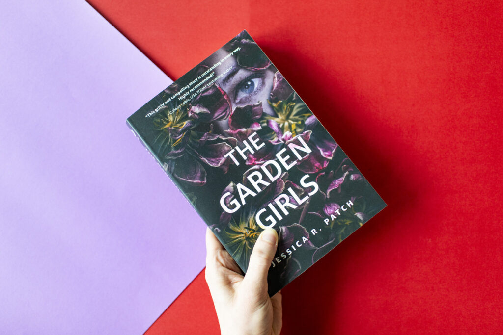 An image of a hand holding up a print copy of THE GARDEN GIRLS by Jessica R. Patch over a red and lavender background.