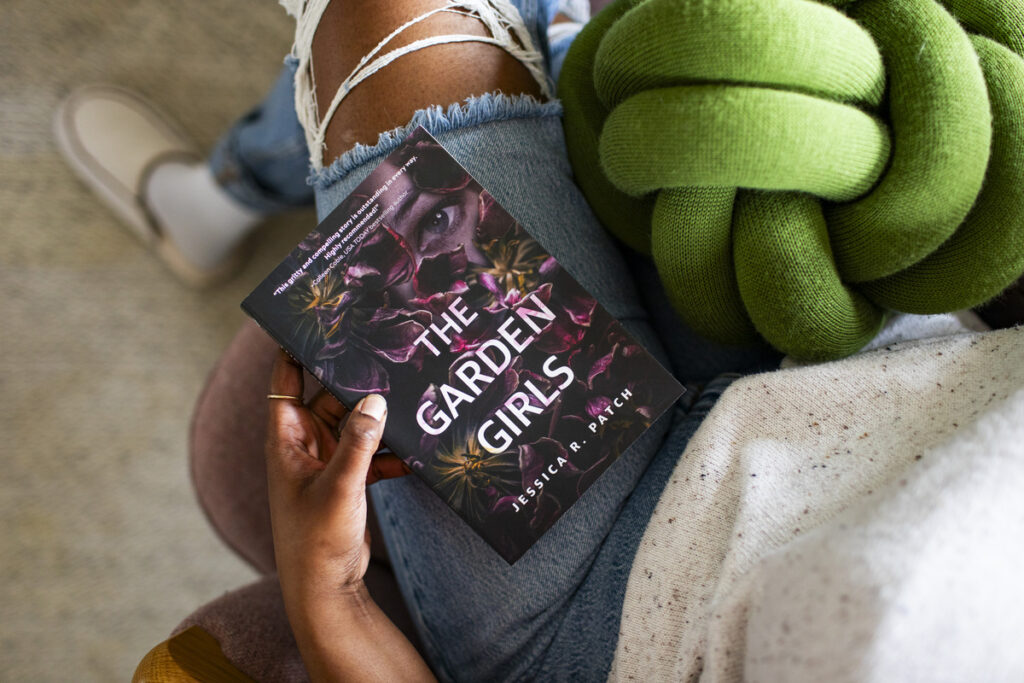 An image of a woman's lap. She is sitting in a green chair and wearing jeans, with a copy of The Garden Girls by Jessica R. Patch resting on her leg.
