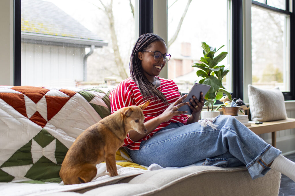 Image of a woman sitting on a couch with her dog, reading from an ereader and smiling.