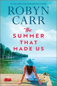 Cover image for The Summer That Made Us by Robyn Carr, featuring a woman sitting on a dock looking over a lake. There are trees in the distance. The woman is wearing a large blue sunhat.